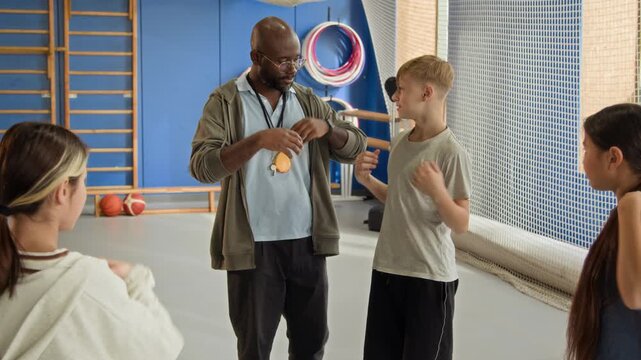 Waist up shot of Black man teaching PE class explaining shoulder rolls exercise to group of children and correcting technique