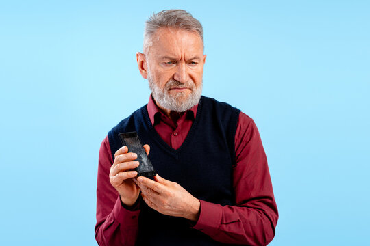 Sad unhappy senior man wearing casual clothes holding broken mobile phone with cracked display