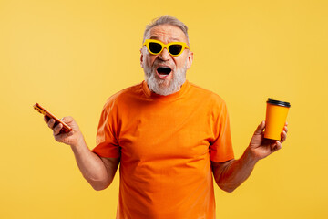 Happy senior man wearing sunglasses holding mobile phone with open mouth on yellow background