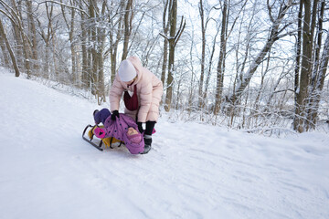 Mother in pink coat supporting her daughter on a sled during winter walk in forest