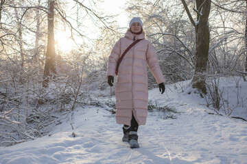 Young woman in long pink down jacket walking on snowy path in winter park