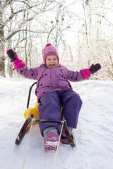 Excited little girl raising arms with joy while sitting on sled in sunny winter park