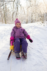 Happy little girl sitting on yellow sled and smiling during winter walk in snowy park