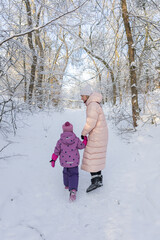 Young mother and little daughter walking hand in hand on snowy path in winter forest