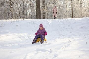 Close up of little girl riding yellow sled down snowy hill in bright winter sunlight