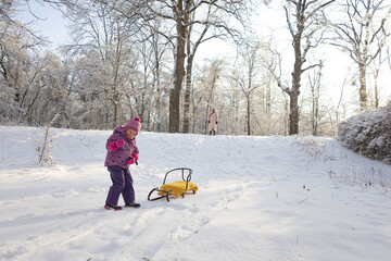Little girl standing in snow with yellow sled against bright winter sun in forest park