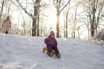 Little girl in purple suit sledding down snowy hill on yellow sled in sunny winter forest