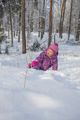 Little girl in warm winter clothes sitting in snowbank and looking away in beautiful winter forest