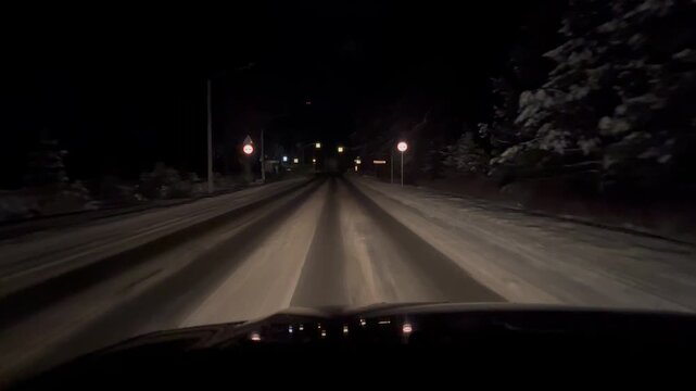 A snowy road at night with faint streetlights illuminating the path. The scene is dark and quiet, showcasing winter conditions.