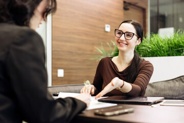 Smiling woman with glasses is engaged in a conversation with another person at a modern office table, documents and devices visible in a professional setting with greenery