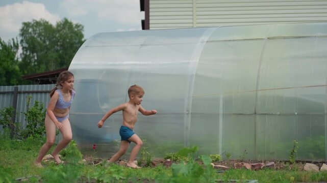 Children Running Near Plastic Greenhouse, Playful Group Sprint Across Garden Beds Under Bright Summer Sky, Candid Energy And Shared Discovery As Kids Chase And Explore Outdoor Plants