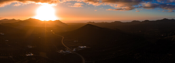 Aerial view of Lanzarote at sunset shows La Geria vineyards, conical lava hills, and a winding road by whitewashed buildings, with amber rays and jagged ridges.
