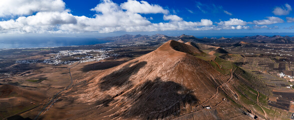 Aerial view of Lanzarote, Canary Islands, showing La Geria vineyards, Timanfaya craters, Arrecife, lava fields, ochre cones, winding roads, and white villages under cumulus clouds. © Aerial Film Studio