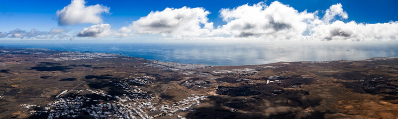 Aerial view of Lanzarote southeast coast shows Arrecife, Puerto del Carmen, dark lava plains, cratered terrain, winding roads, and a distant ferry under soft cloud cover. © Aerial Film Studio