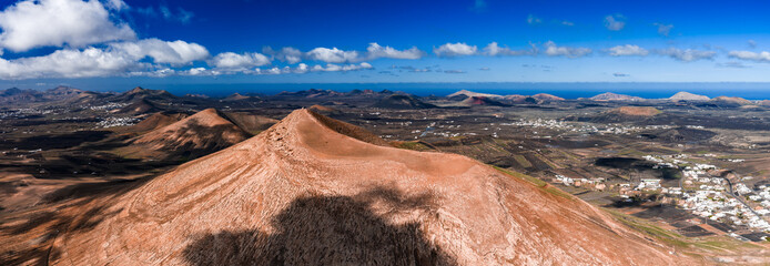 Aerial view shows an ochre volcanic cone in Lanzarote, Canary Islands, with La Geria vineyards, whitewashed villages, and the Atlantic horizon under midday light. © Aerial Film Studio