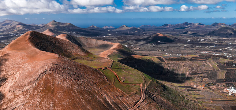Aerial view shows Lanzarote, Canary Islands, with ochre caldera ridges, La Geria vine pits and stone walls, white houses, and conical volcanoes under patchy daylight.