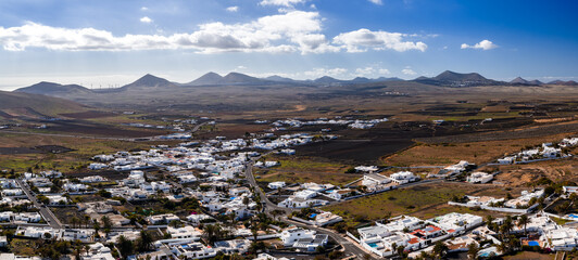 Aerial daytime view of a whitewashed town on black lava fields near La Geria, Lanzarote. Conical lava hills, craters, stone walled plots, and wind turbines are visible. © Aerial Film Studio