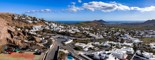 White Canarian houses span volcanic hills near Teguise and La Geria, palm lined streets and terraced lava slopes, midday light, green patches, cobalt sea visible. © Aerial Film Studio