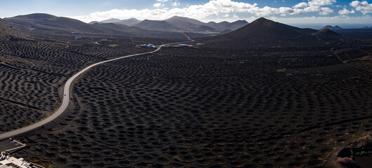 Aerial view of La Geria vineyards in Lanzarote shows circular hollows in black ash, a curving road, whitewashed buildings, and Timanfaya cones under scattered clouds. © Aerial Film Studio