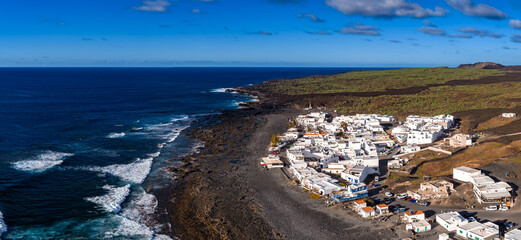 Aerial view of a Lanzarote coastal village in the Canary Islands, whitewashed houses by black lava shore, Atlantic horizon, midday sun, clear sky, basalt flows, tide pools.