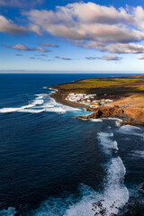 Aerial view shows El Golfo with whitewashed houses by Timanfaya lava fields and a dark volcanic shore. Atlantic swells line black coves under soft afternoon light. © Aerial Film Studio