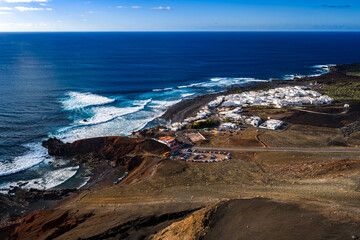 Aerial view of El Golfo, Lanzarote, Canary Islands, with lava cliffs, curved bay, green lagoon area, whitewashed village, restaurants, car park, surf, and midday light. © Aerial Film Studio