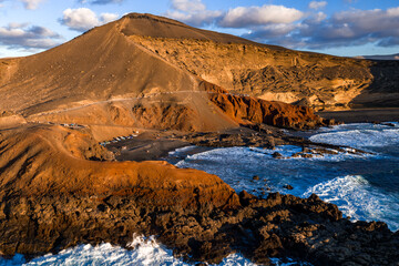 Aerial view of Lanzarote at El Golfo near Charco Verde. Rust cliffs meet black lava rocks and surf. A coastal path and parked cars show visitors in late afternoon light.