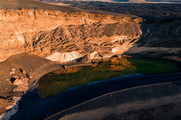 Aerial view of Charco Verde on Lanzarote, Canary Islands, with emerald algae lake, ebony lava sand, ochre cliffs, crescent beach, near Timanfaya in late day light.