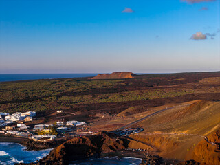 Rust colored volcanic cliffs, black basalt, and ochre cones frame a whitewashed village by a cove near El Golfo and Charco Verde in Lanzarote, with walkers on a road. © Aerial Film Studio