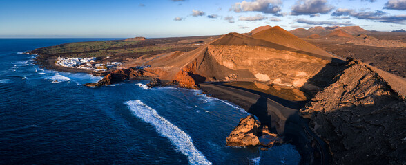 Aerial view shows El Golfo amphitheater, Charco de los Clicos area, and black sand on Lanzarote. Ocean swells curl as a village sits left, lava fields and tuff cliffs right. © Aerial Film Studio