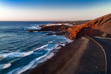 Aerial view of Lanzarote near El Golfo shows rust volcanic cliffs, black sand, and turquoise surf. White houses sit above rocky coves by Charco de los Clicos. © Aerial Film Studio