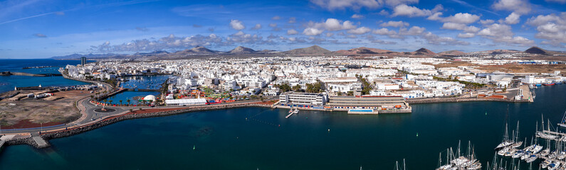 Wide aerial view of Arrecife, Lanzarote, with whitewashed buildings by the harbor and marinas, curving promenade, moored boats, and volcanic cones under crisp midday light. © Aerial Film Studio