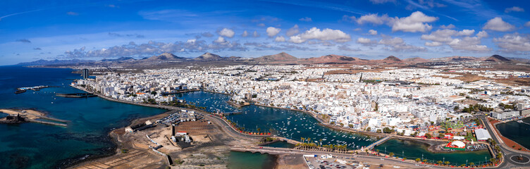 Aerial view of Arrecife, Lanzarote, with Charco de San Gines, marina, Gran Hotel, arched bridges, palm promenades, boats, piers, and volcanic cones under midday light. © Aerial Film Studio