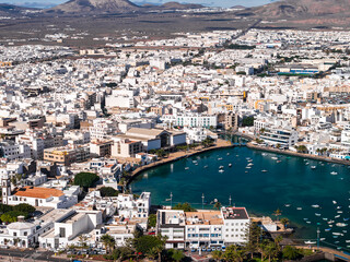 Aerial midday view of Arrecife, Lanzarote, with Charco de San Gines, small boats, a small arched bridge, palm promenades, and volcanic hills and lava fields. © Aerial Film Studio