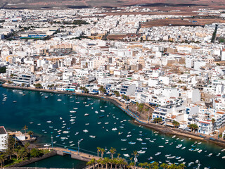 Aerial view of Arrecife, Lanzarote shows whitewashed buildings by Charco de San Gines with small boats, palm promenade, low bridges, and inland russet volcanic terrain at midday. © Aerial Film Studio