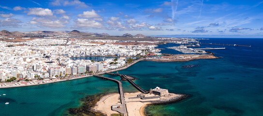 Aerial view of Arrecife, Lanzarote shows whitewashed city, Castillo de San Gabriel, causeway, marina, port, cobalt Atlantic, volcanic hills in crisp midday light. © Aerial Film Studio