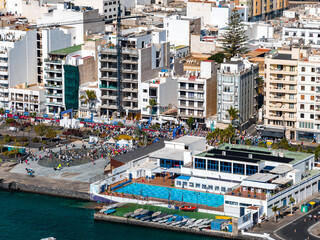 Midday aerial shows Arrecife, Lanzarote waterfront, whitewashed blocks, palm trees, seaside pool lanes, and marina as crowds line the marathon route along the harbor. © Aerial Film Studio