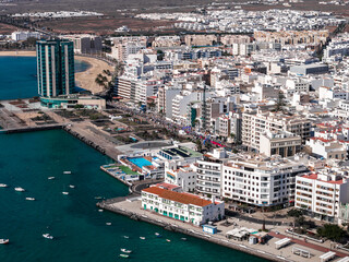 Aerial view of Arrecife, Lanzarote, shows seafront promenade, small marina, Playa del Reducto, and Gran Hotel and Spa. Crowds and banners suggest 2025 marathon route. © Aerial Film Studio