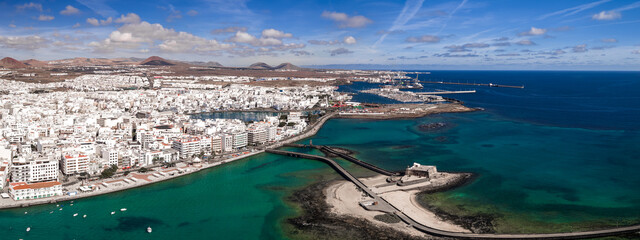 Aerial view of Arrecife, Lanzarote, with whitewashed buildings by emerald waters. Castillo de San Gabriel, twin bridges, marina, and volcanic cones appear at midday. © Aerial Film Studio
