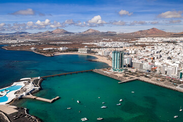 Aerial view of Arrecife, Lanzarote, with Playa del Reducto, Arrecife Gran Hotel and Spa, a seafront promenade, small boats, whitewashed buildings, and volcanic peaks. © Aerial Film Studio