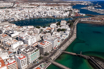 Obraz premium Aerial view of Arrecife, Lanzarote shows Charco de San Gines with small boats, a promenade, the church of San Gines, a stone causeway, breakwater, and the working harbor.