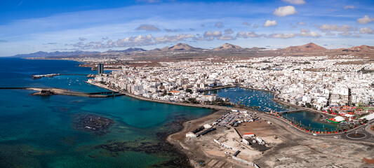 Aerial view of Arrecife, Lanzarote shows turquoise Atlantic coast, El Charco de San Gines with boats, Gran Hotel Arrecife, Castillo de San Gabriel, and inland volcanic cones.