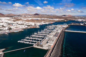 Aerial view shows Arrecife marina and port in Lanzarote with sailboats, breakwaters, whitewashed buildings, volcanic hills, turquoise Atlantic, piers, and seawalls in daylight. © Aerial Film Studio
