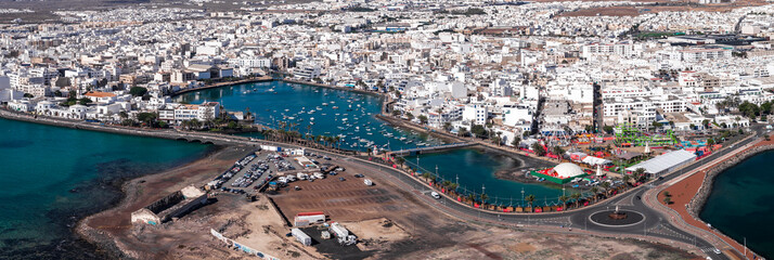 Aerial view of Arrecife, Lanzarote, showing Charco de San Gines, arched bridges, boats, palm promenades, roundabouts, Ferris wheel, tents, rocks, and cubic buildings at midday. © Aerial Film Studio
