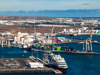 Aerial view of the Port of Arrecife in Lanzarote shows cranes, container stacks, fuel tanks, a docked luxury cruise yacht, industrial silos, and a wind turbine at midday. © Aerial Film Studio