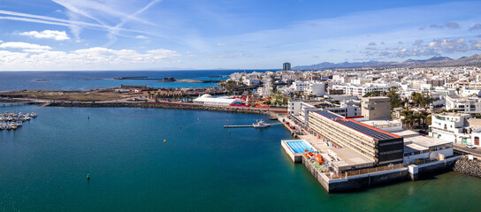 Aerial view of Arrecife, Lanzarote, shows harbor, Castillo de San Gabriel, Gran Hotel Arrecife, marina, seaside pools, solar topped roofs, and volcanic ridges in daylight. © Aerial Film Studio