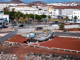 An old wooden sailboat with twin masts sits dry docked by a waterfront roundabout in Arrecife, Lanzarote, with whitewashed buildings, palm trees, and volcanic hills. © Aerial Film Studio