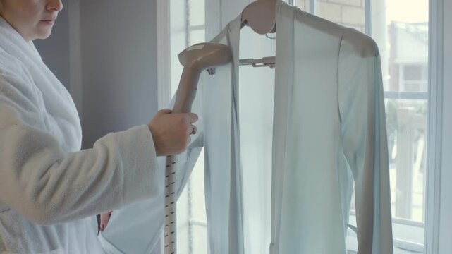Anonymous woman in robe steaming a silk cardigan with a steamer in a cozy bedroom. The models face is not visible. Focus on hands and action.