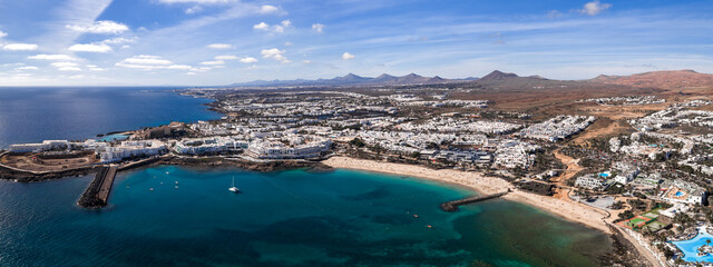 Aerial view of Costa Teguise in Lanzarote shows turquoise bays, golden sand, whitewashed buildings, volcanic cones, protected coves, a marina, pools, and calm seas at daytime. © Aerial Film Studio