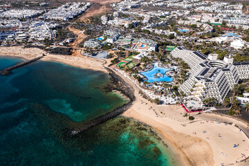 Aerial scene shows Playa de las Cucharas with lava rock breakwaters, Hotel Melia Salinas with lagoon pools, palm promenades, tennis courts, and whitewashed buildings at midday. © Aerial Film Studio